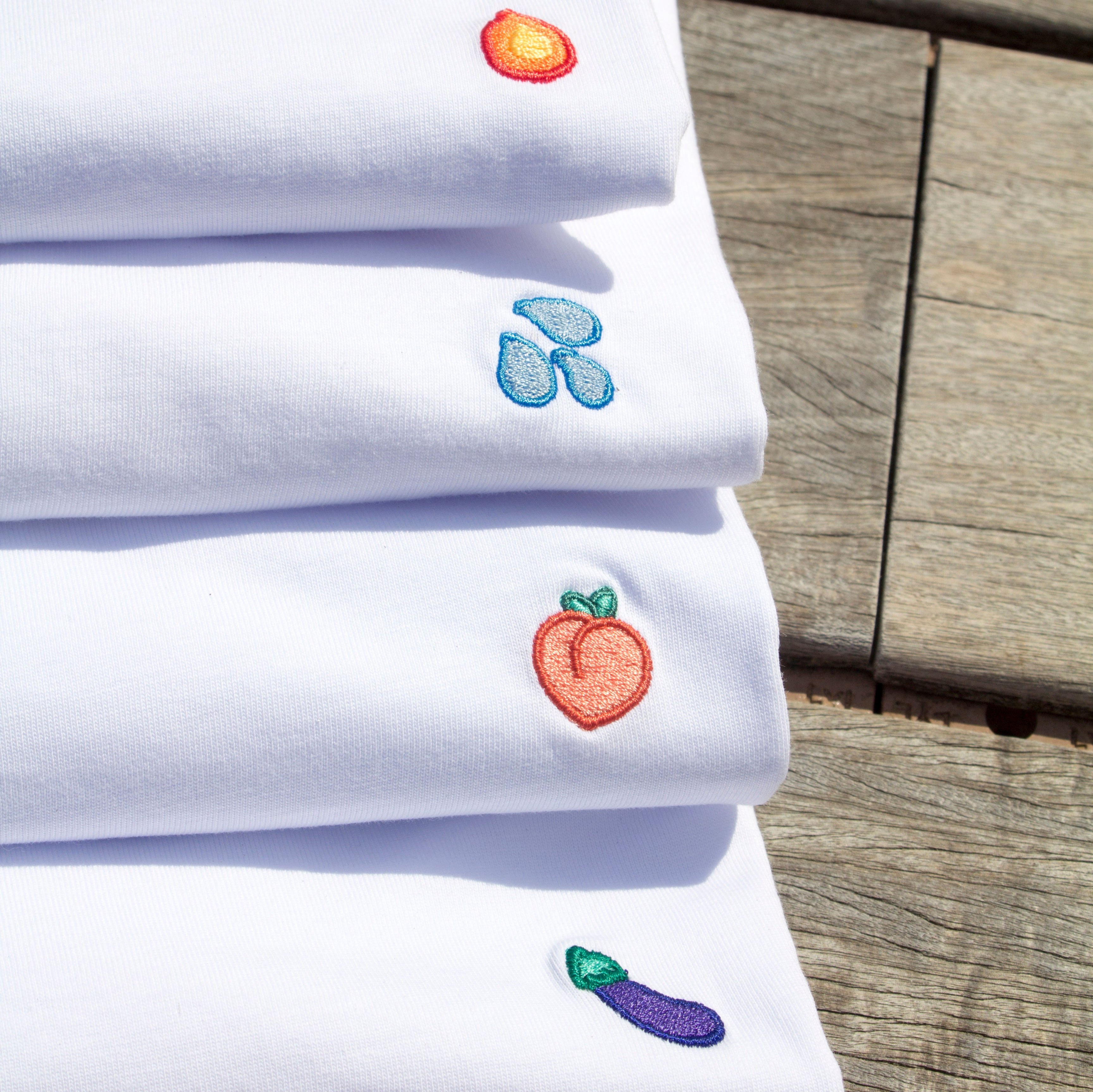 Pile of four folded Out Now! t-shirts featuring eggplant, peach, water drops, and flame patterns, showcasing bold and playful LGBTQ+ designs.