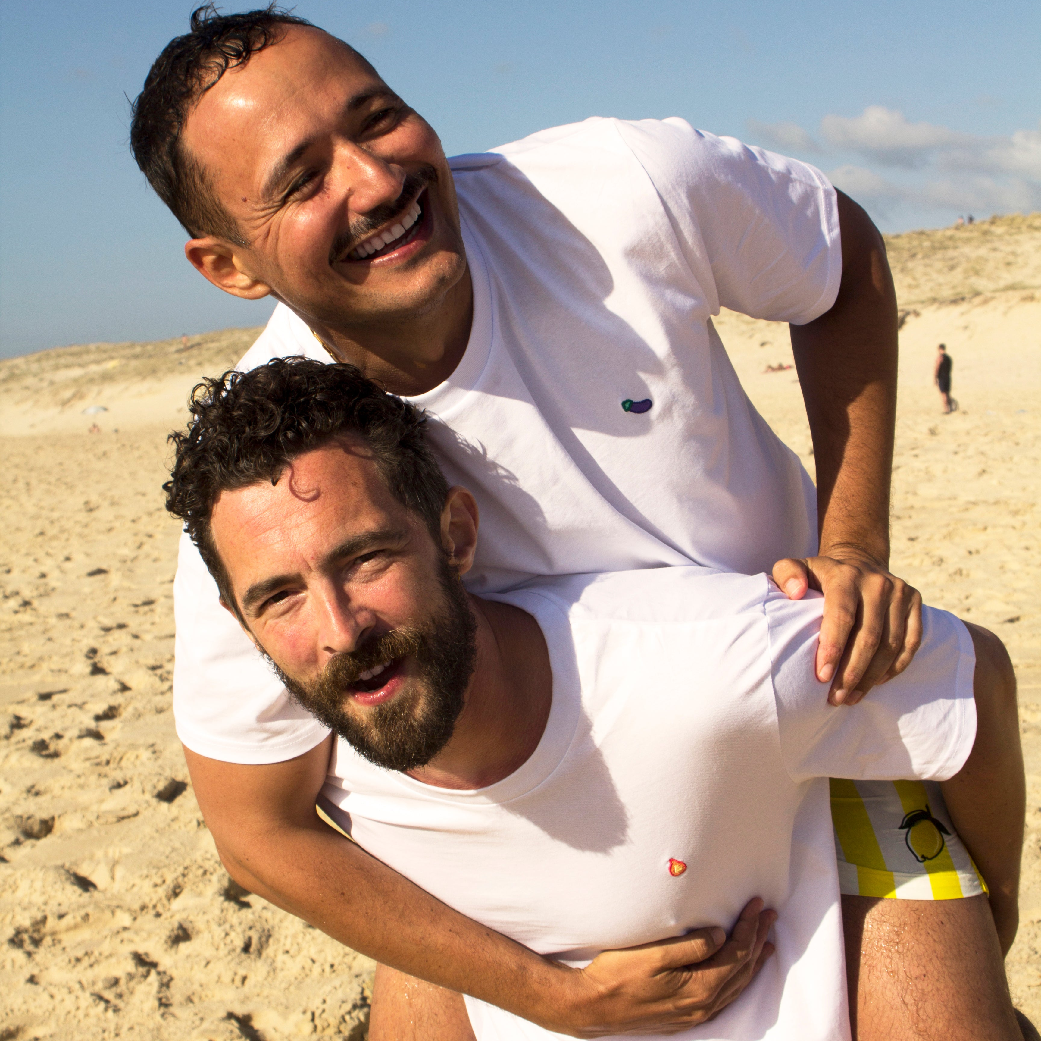 Two male models on the beach wearing Out Now! flame and eggplant t-shirts, embodying the brand's values of diversity, connection, and LGBTQ+ pride.