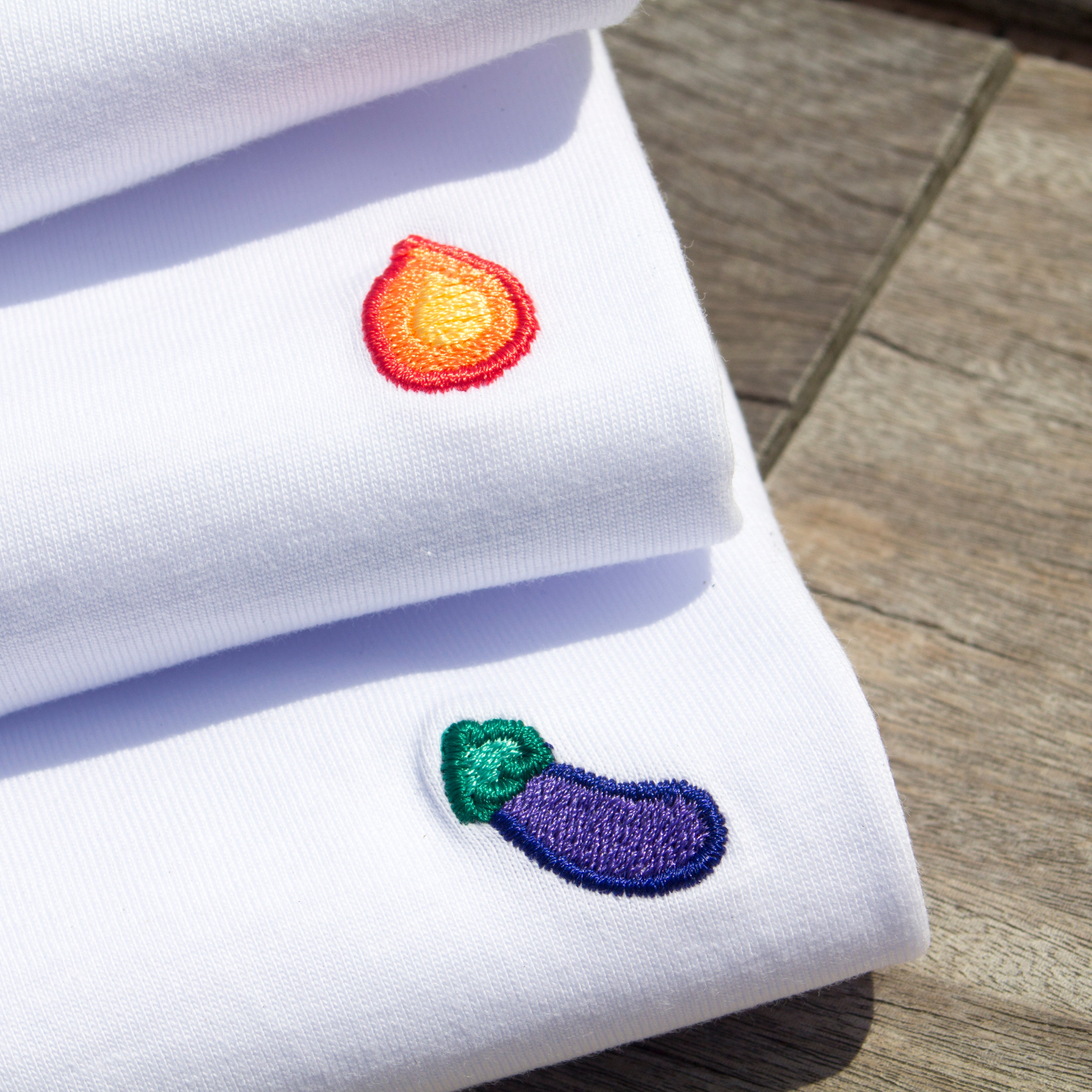 Detailed view of flame and eggplant embroidery on stacked white Out Now! t-shirts, highlighting fine stitching and vibrant designs, reflecting LGBTQ+ pride and identity.