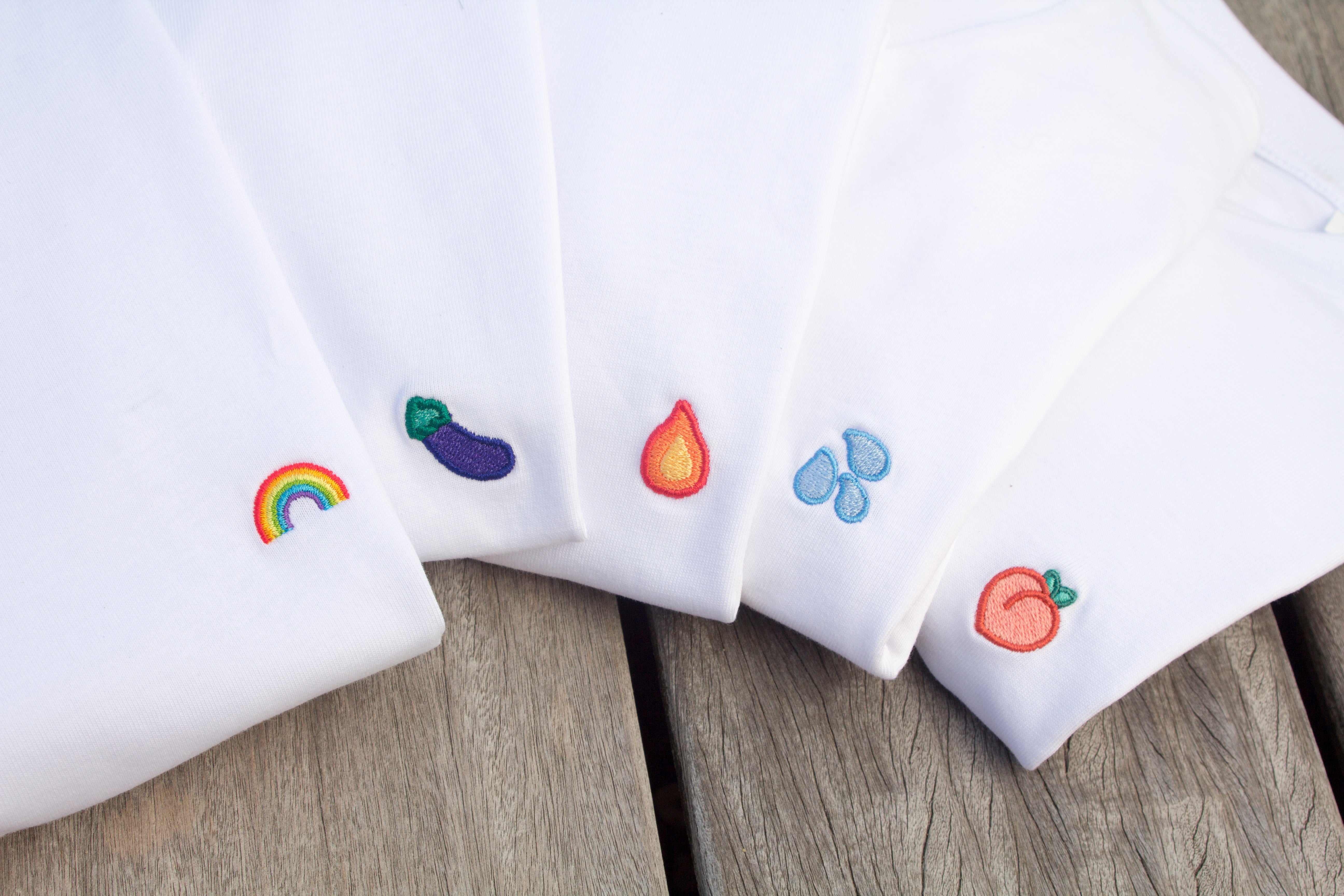 Five Out Now! t-shirts fanned out, featuring vibrant rainbow, eggplant, peach, water drops, and flame patterns, celebrating LGBTQ+ pride through bold, playful designs.