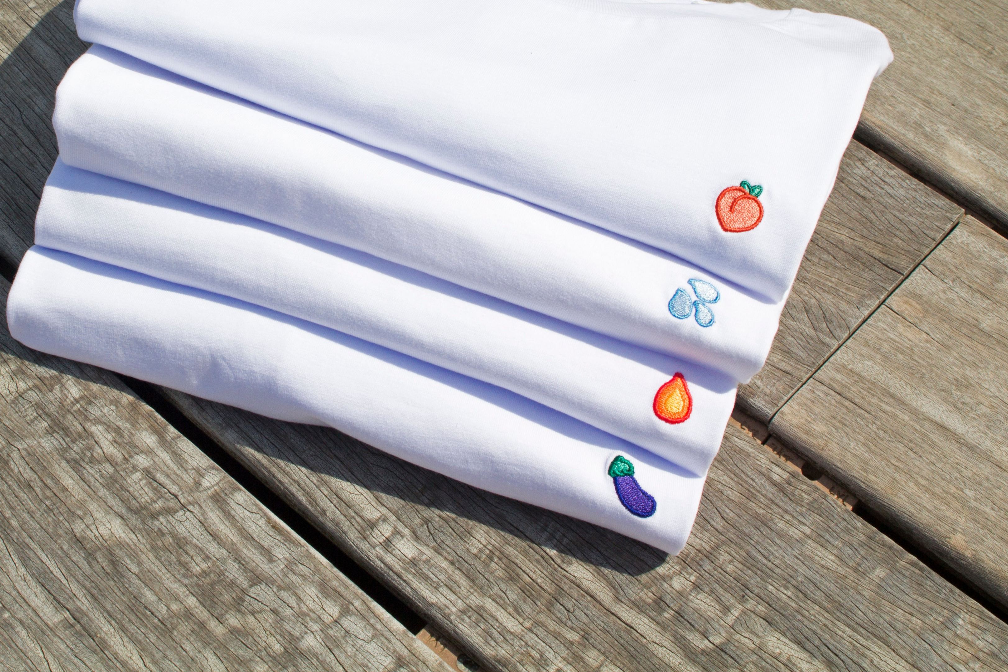 Four folded Out Now! t-shirts stacked, featuring vibrant eggplant, peach, water drops, and flame patterns, celebrating LGBTQ+ pride through bold and playful designs.