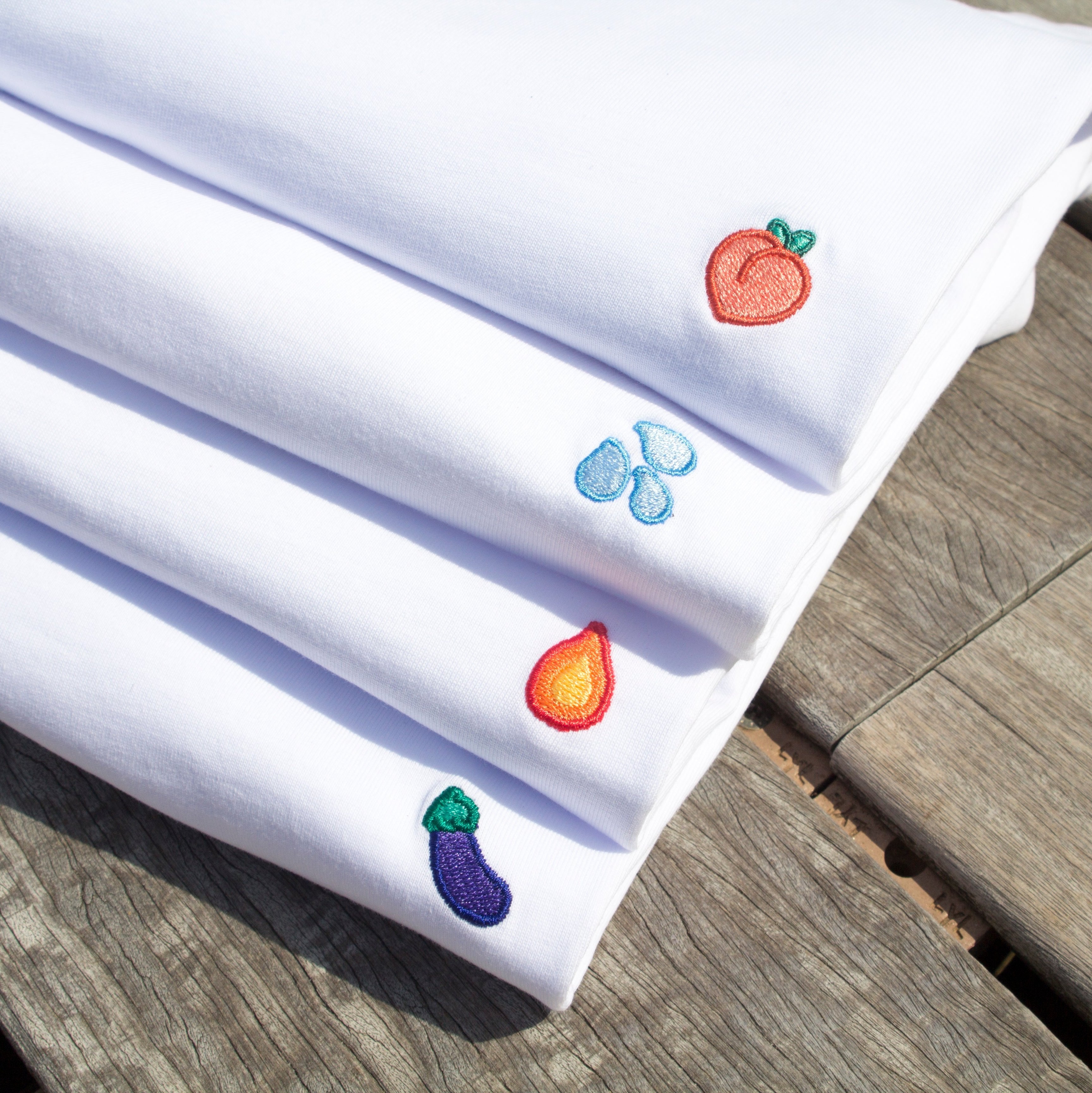 Stack of four folded Out Now! t-shirts featuring bold patterns—eggplant, peach, water drops, and flame—highlighting playful designs inspired by LGBTQ+ pride and identity.